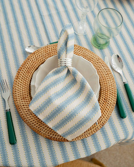 Table setting with a striped napkin, woven plate, and cutlery on a striped tablecloth.