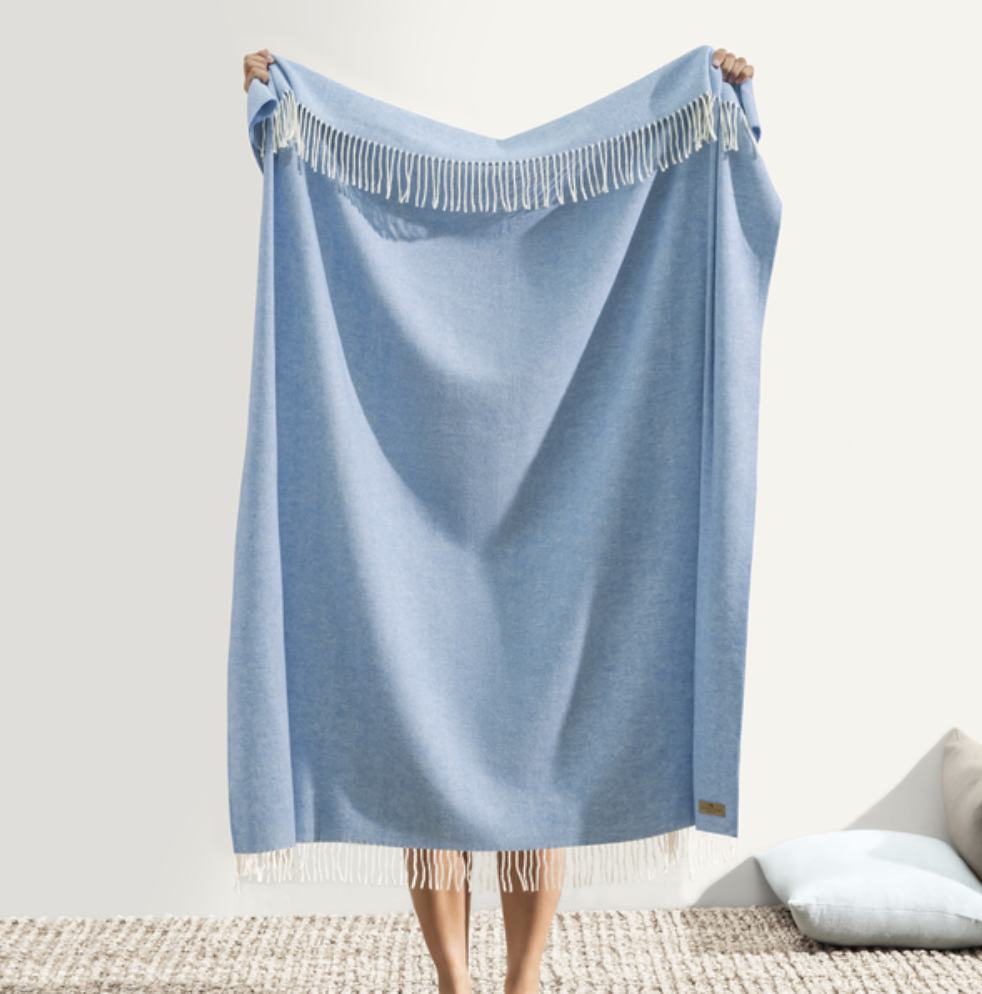 Person holding a light blue throw blanket with fringe on a neutral background
