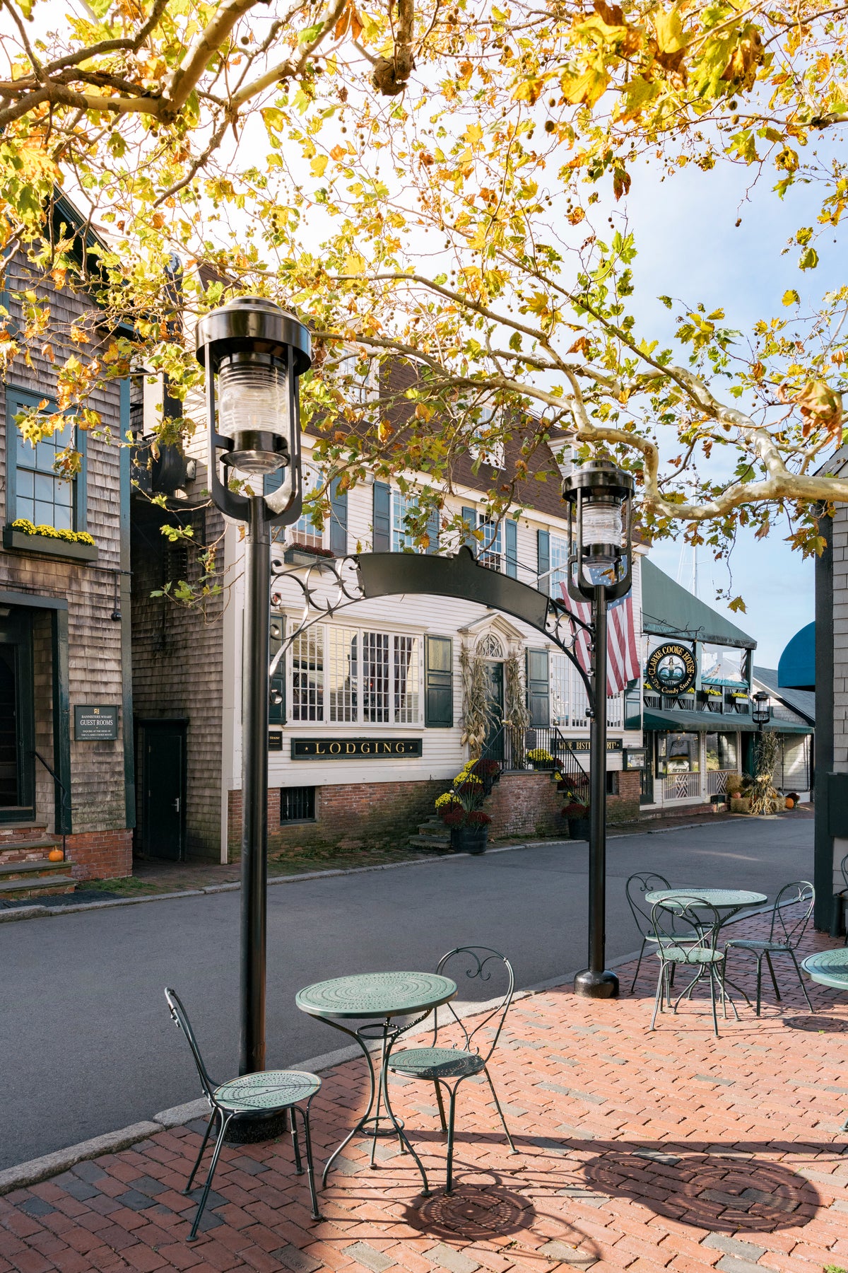 Outdoor street scene with brick tables and chairs under black metal lamp posts, framed by autumn trees with yellow leaves. Historic buildings line the street, including one with a visible 'LODGING' sign.