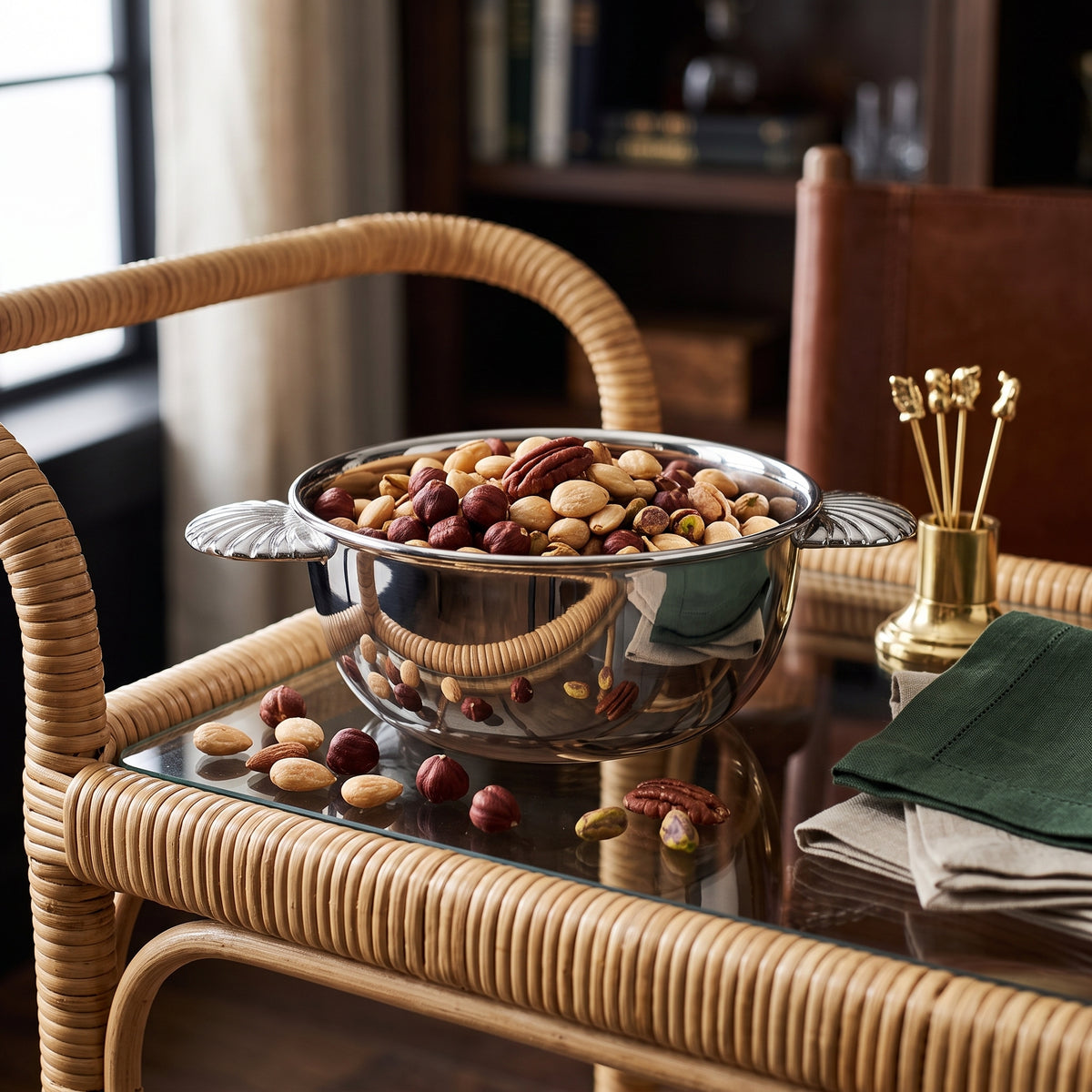 A polished stainless steel bowl filled with assorted nuts and dried fruits, styled on a rattan bar cart in a warm, elegant room setting.