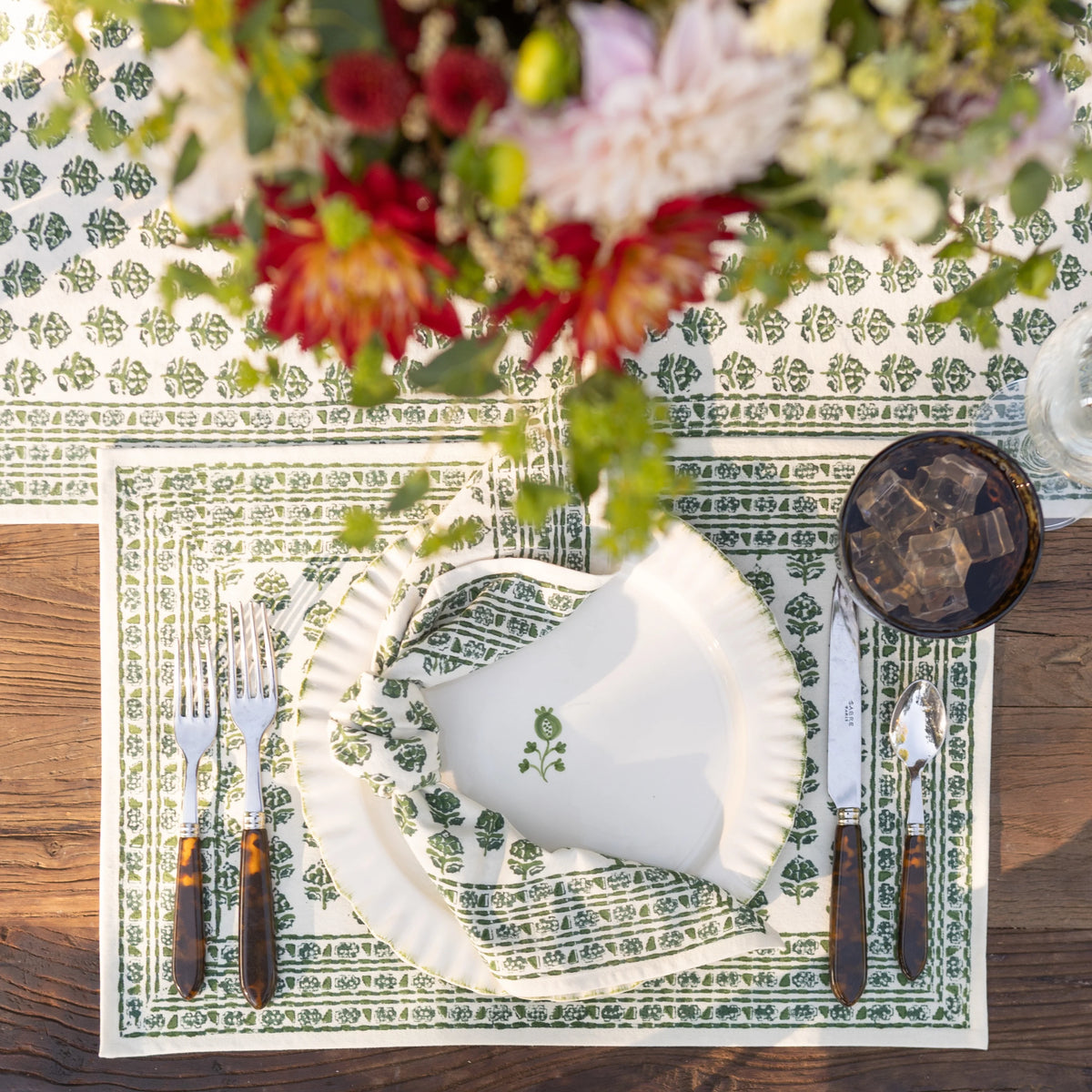 A styled table setting featuring a white paper plate and matching napkin with a green botanical border, placed on a green and white patterned placemat over a rustic wooden table. Silverware with dark wood handles, a glass of iced coffee, and a blurred floral arrangement are also visible.