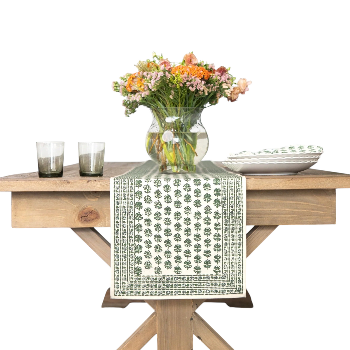 A green and white patterned table runner draped over a light wood table, styled with a glass vase of orange and pink flowers, two gray candles, and white plates in a bright room.