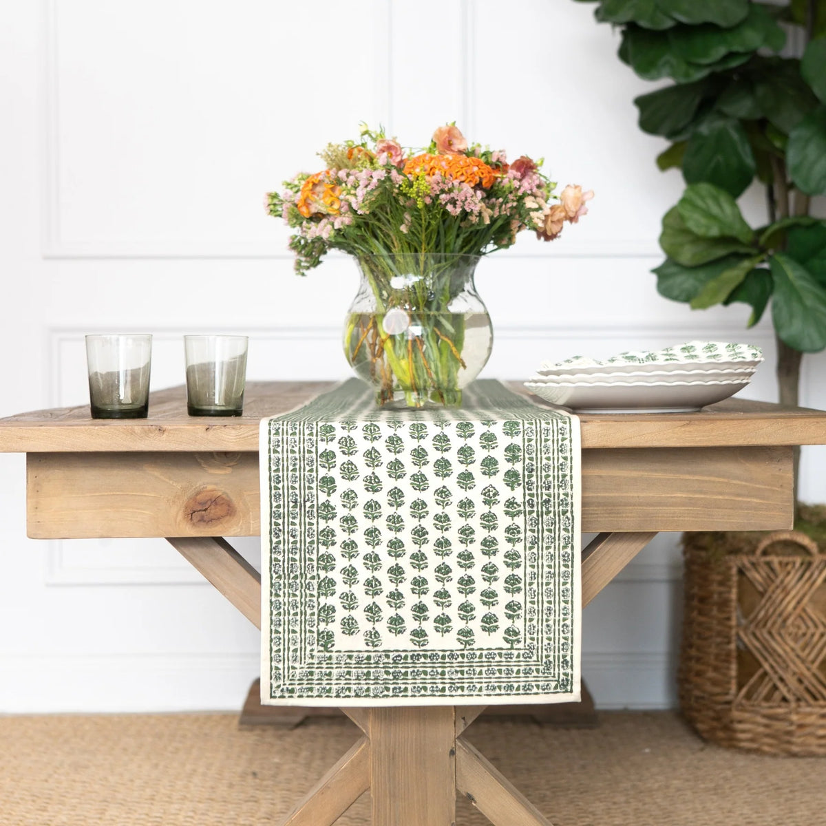 A green and white patterned table runner draped over a light wood table, styled with a glass vase of orange and pink flowers, two gray candles, and white plates in a bright room.