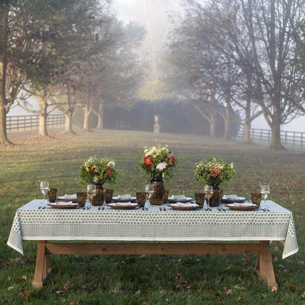 A rustic outdoor dining table set with a white patterned tablecloth, plates, glasses, and three floral centerpieces, photographed in a misty park with trees and grass in the background.