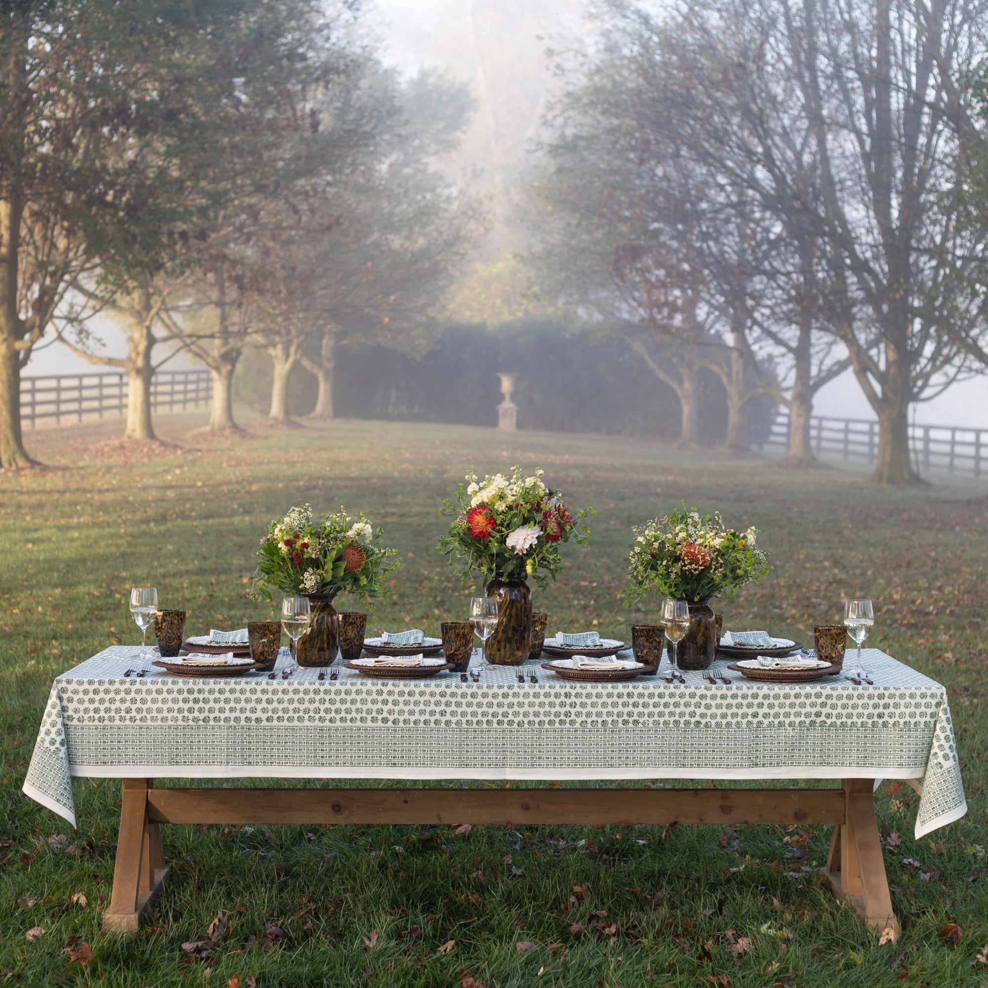 A rustic outdoor dining table set with a white patterned tablecloth, plates, glasses, and three floral centerpieces, photographed in a misty park with trees and grass in the background.