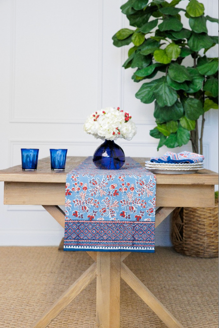 Decorative table runner with floral pattern on a wooden table with blue glasses and a vase.