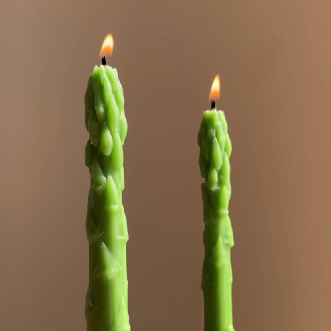 Two green asparagus-shaped candles with flames on a beige background