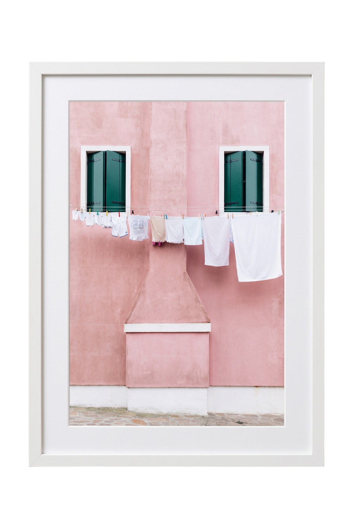 A framed art print featuring a pink stucco building facade with two dark green shuttered windows and white laundry hanging on a clothesline, set in a white frame against a white background.