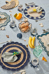 Table setting with woven placemats, striped tablecloth, and various items on a blue and white striped tablecloth.