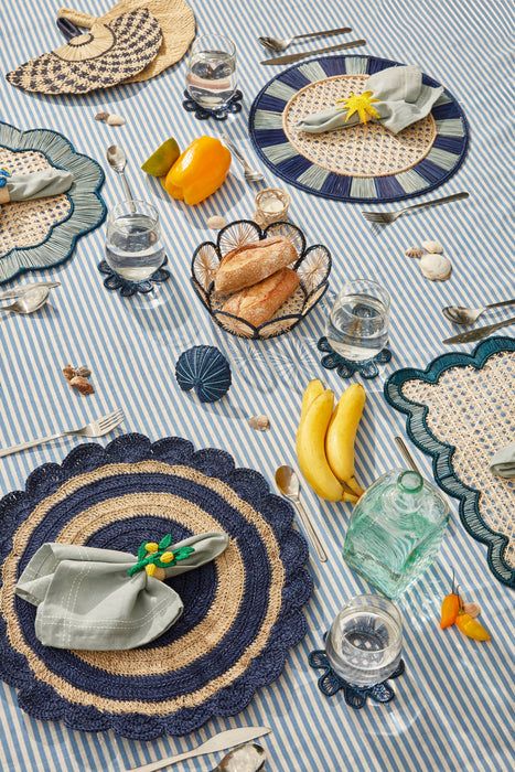 Table setting with woven placemats, striped tablecloth, and various items on a blue and white striped tablecloth.