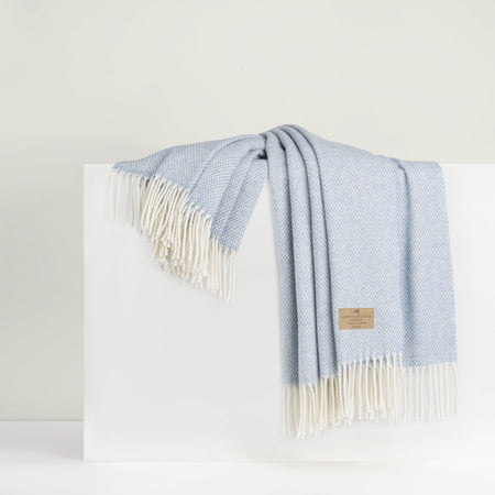 A light blue basket weave cashmere and lambswool throw with fringes displayed on a white platform.