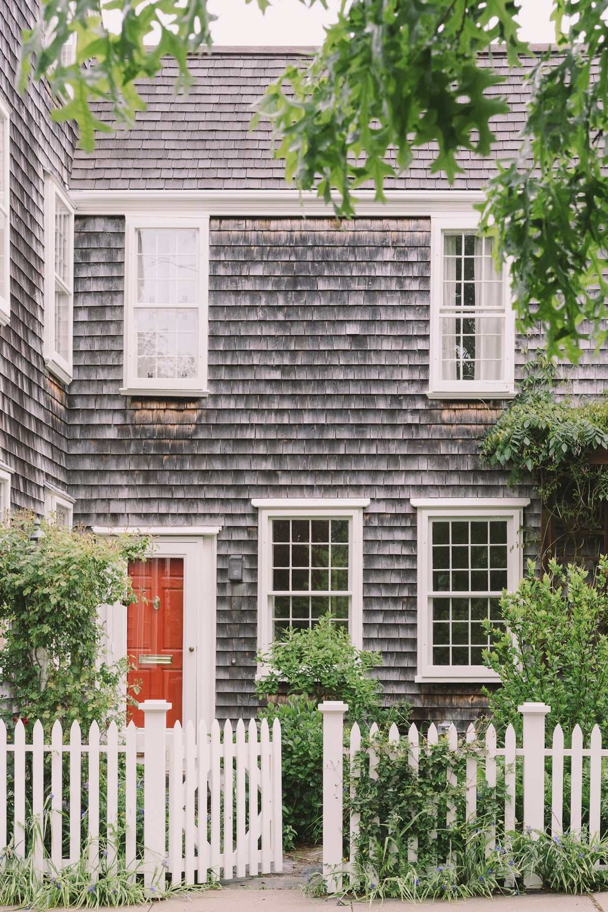 Traditional house with gray shingles, white windows, and a red door, surrounded by greenery.