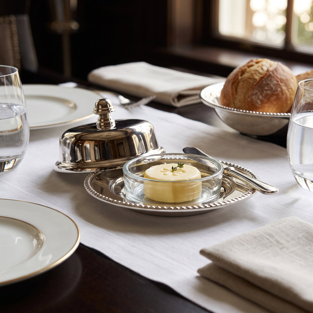 A silver butter dish with a glass bowl of butter on a decorative silver plate, set on a white tablecloth in a dining setting with plates, bread, and water glasses.