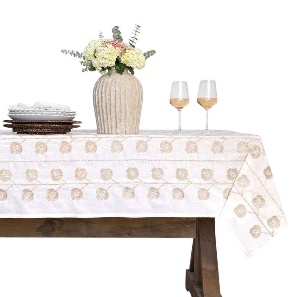 An off-white cotton tablecloth with beige floral embroidery, styled on a wooden table with a vase of flowers, plates, and two wine glasses against a white background.