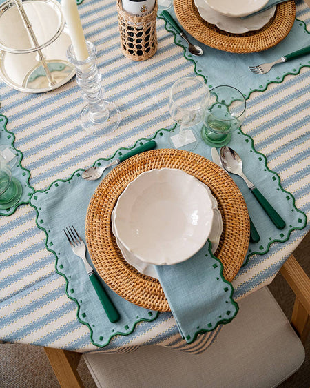 Dining table setting with striped tablecloth, wicker chargers, and green cutlery.