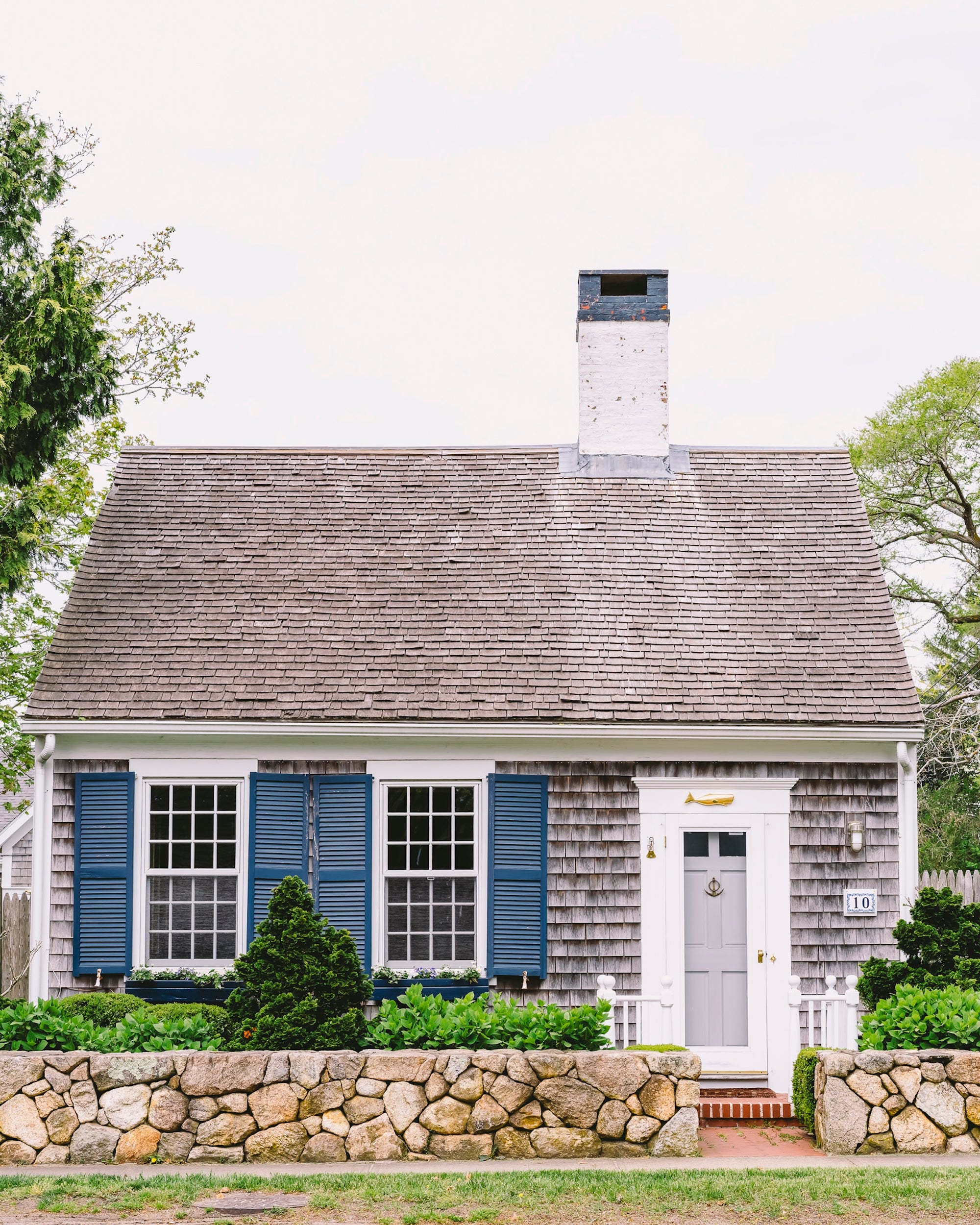 Small house with gray roof and blue shutters, surrounded by greenery.