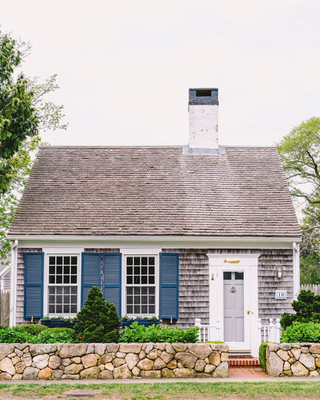 Small house with gray roof and blue shutters, surrounded by greenery.