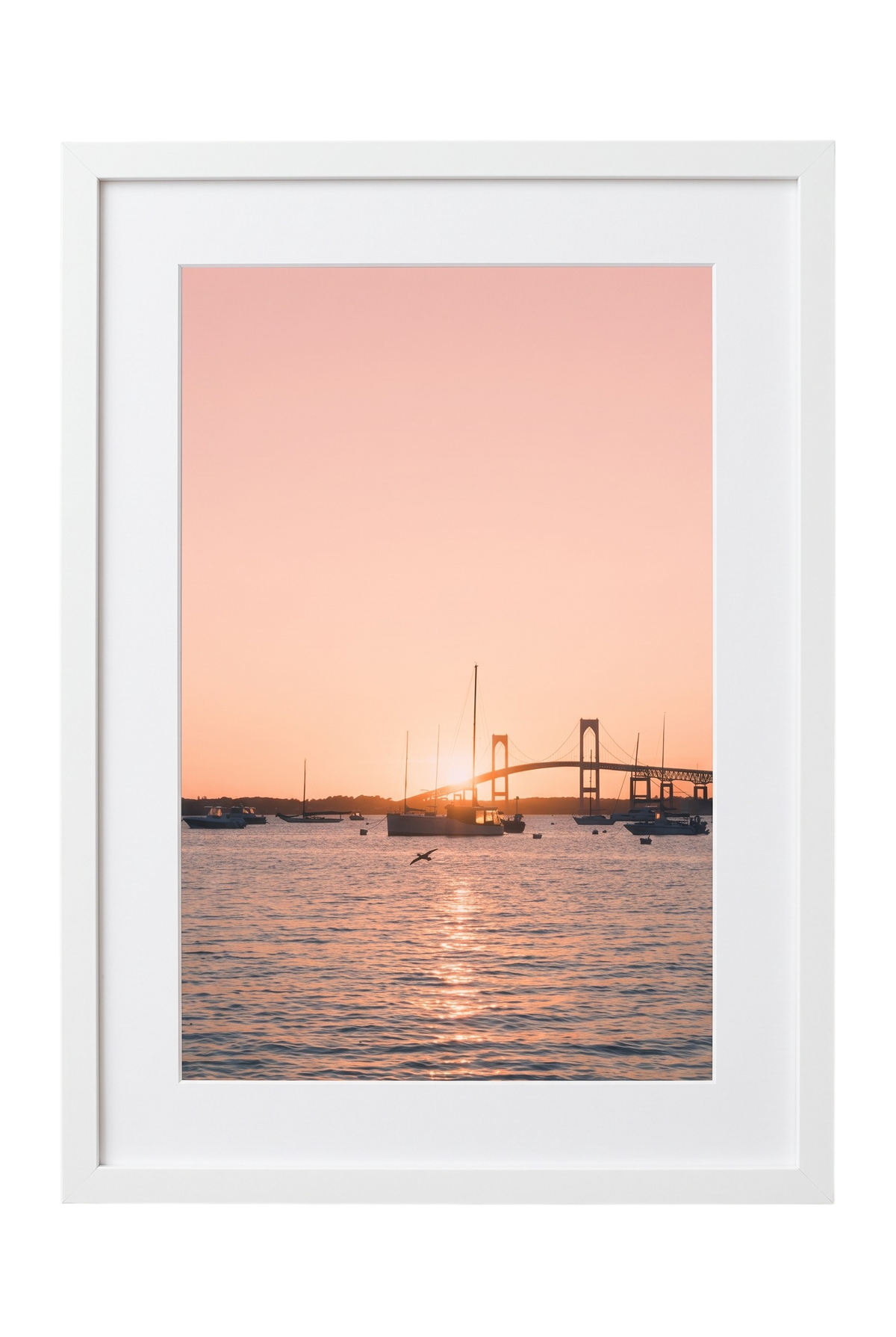 A framed photographic print of a sunset over water with sailboats and a bridge silhouette, presented in a white frame against a white background.