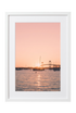 A framed photographic print of a sunset over water with sailboats and a bridge silhouette, presented in a white frame against a white background.