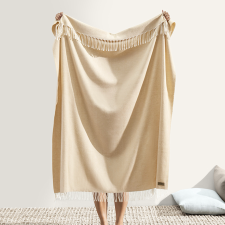 Beige throw blanket with fringe held by a person on a neutral background