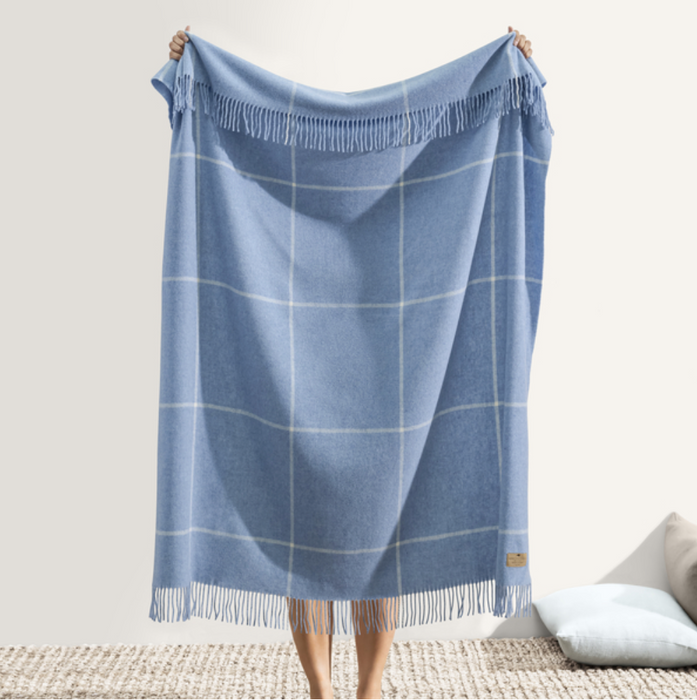 A person holding a sky blue windowpane cashmere throw with a classic checkered pattern and fringed edge detail.