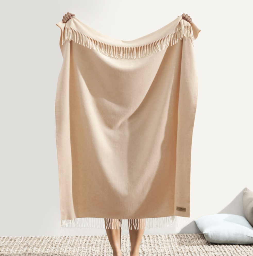 Beige throw blanket with fringes draped over a chair on a light background