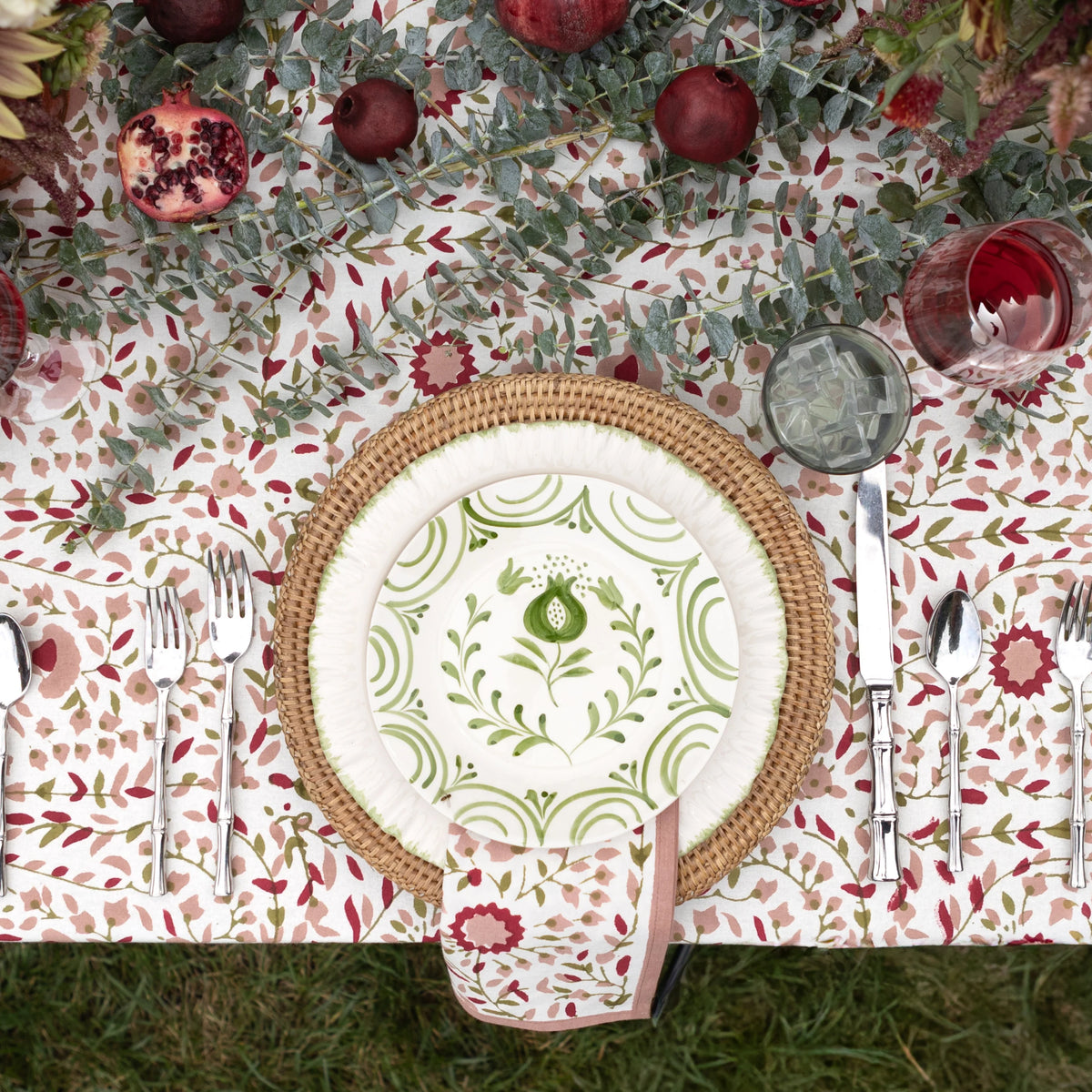 A styled tabletop setting viewed from above, featuring a round woven natural fiber placemat, a white dinner plate with green floral scrollwork and a green poppy motif, a matching folded napkin, silver flatware, a glass of red beverage, and a decorative table runner with greenery, berries, and red fruits on a grassy surface.