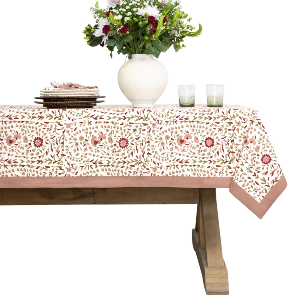 A rectangular tablecloth with a cream background and a repeating floral and leaf pattern in dusty pink, sage green, and terracotta, styled on a wooden dining table with a white vase of flowers, plates, and glasses.
