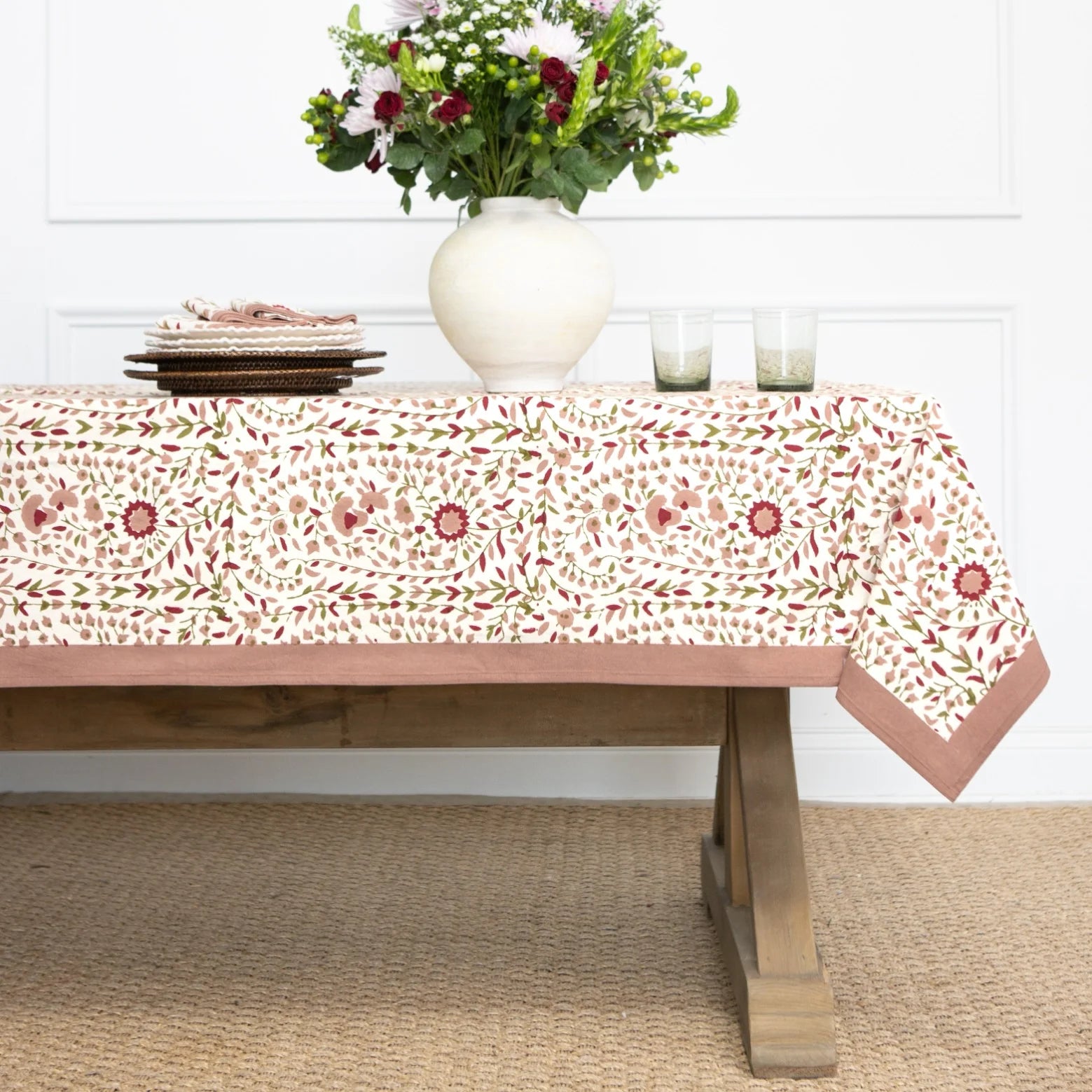 A rectangular tablecloth with a cream background and a repeating floral and leaf pattern in dusty pink, sage green, and terracotta, styled on a wooden dining table with a white vase of flowers, plates, and glasses.