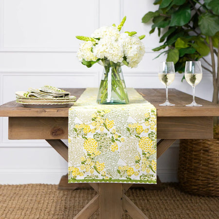 Dining table set with a floral table runner, white flowers, and wine glasses.