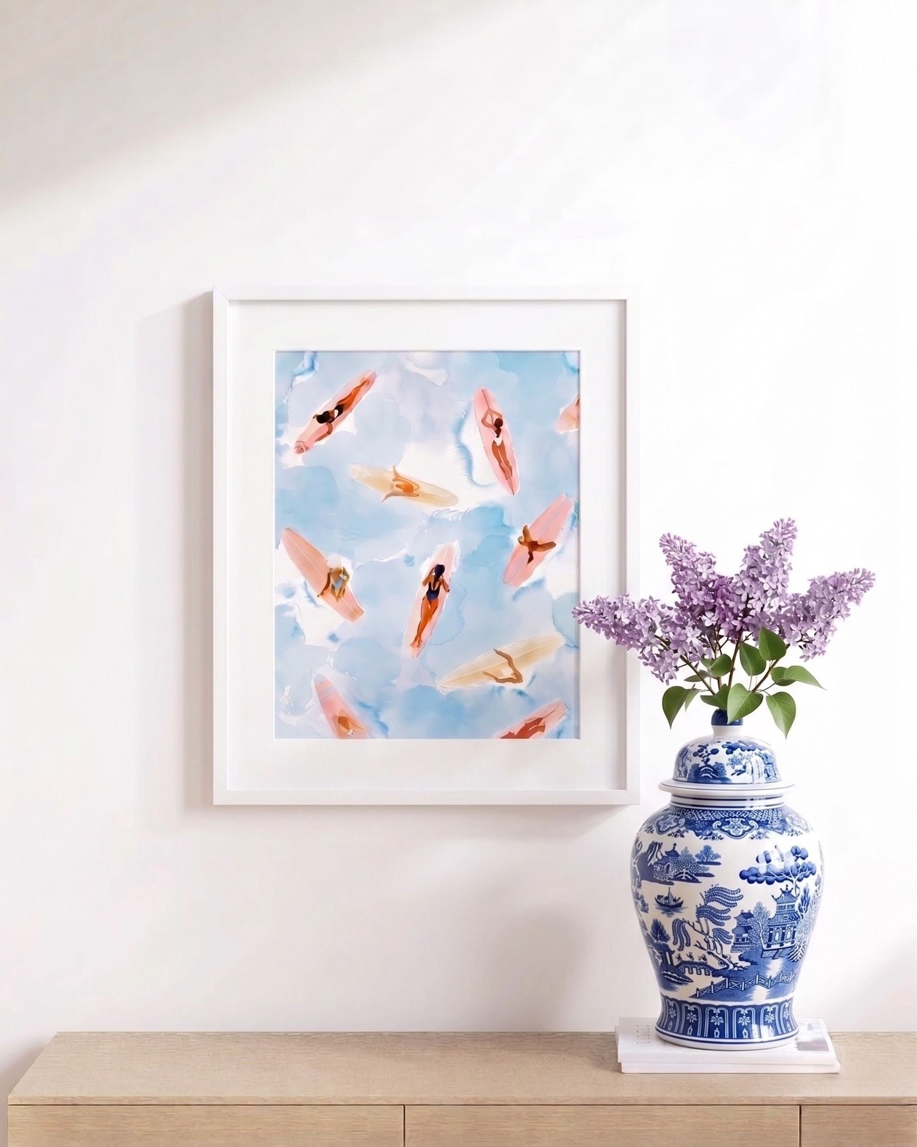 Framed artwork of surfers on a blue sky background with a decorative vase and flowers on a wooden surface.