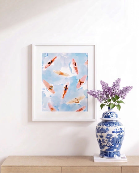 Framed artwork of surfers on a blue sky background with a decorative vase and flowers on a wooden surface.