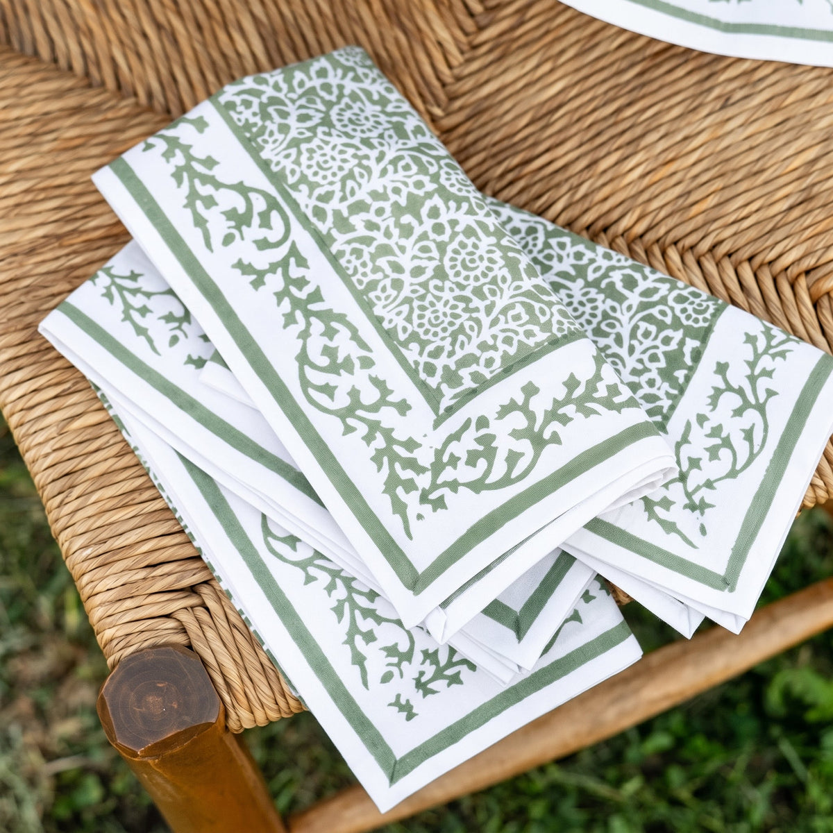 A stack of white paper napkins with a sage green floral damask pattern and border, displayed on a woven wicker chair outdoors.
