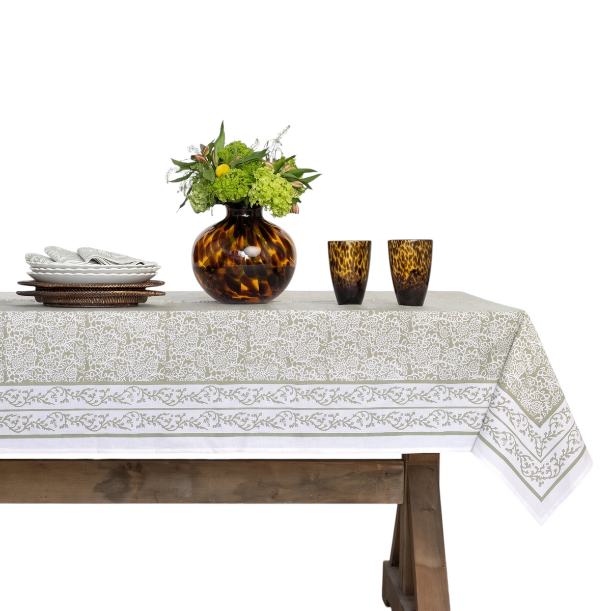 A rectangular tablecloth with a white base and a warm beige floral pattern, draped over a wooden dining table. The table is styled with a dark amber glass vase containing green and yellow flowers, two matching amber and black patterned drinking glasses, and a stack of white plates, set against a white paneled wall.