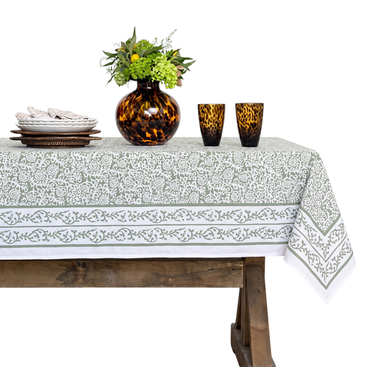 A rectangular tablecloth with a white base and a gray botanical border pattern, styled on a rustic wooden table with a brown glass vase of flowers and two amber patterned drinking glasses against a white paneled wall.