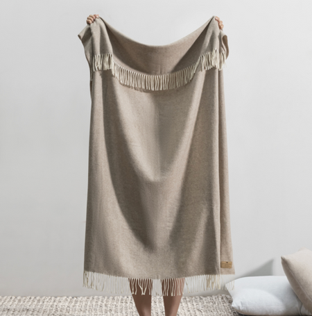 Beige blanket with fringe held by a person against a plain background