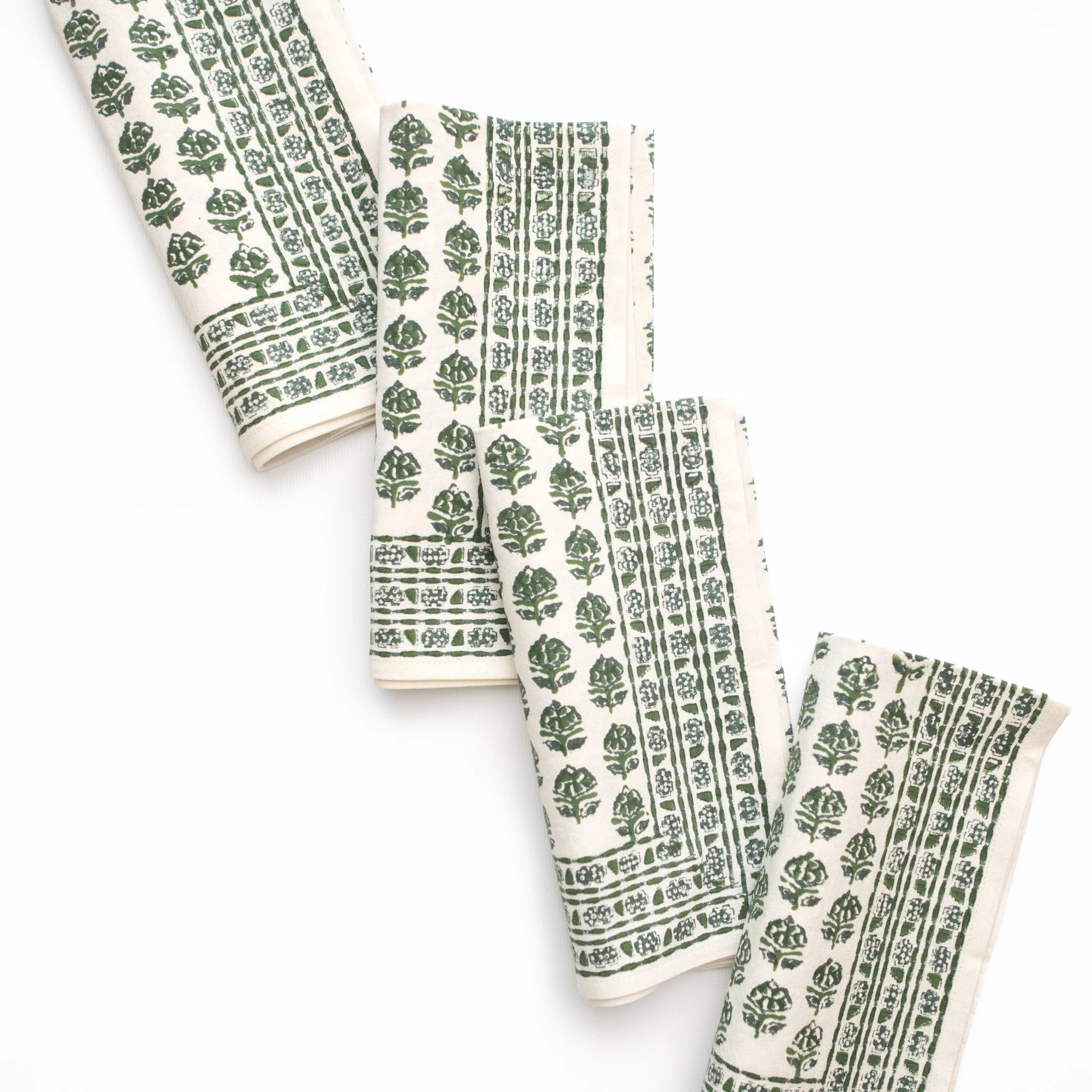 Four folded cotton napkins with a green block-printed leaf and border pattern on an off-white background, arranged diagonally on a white studio background.