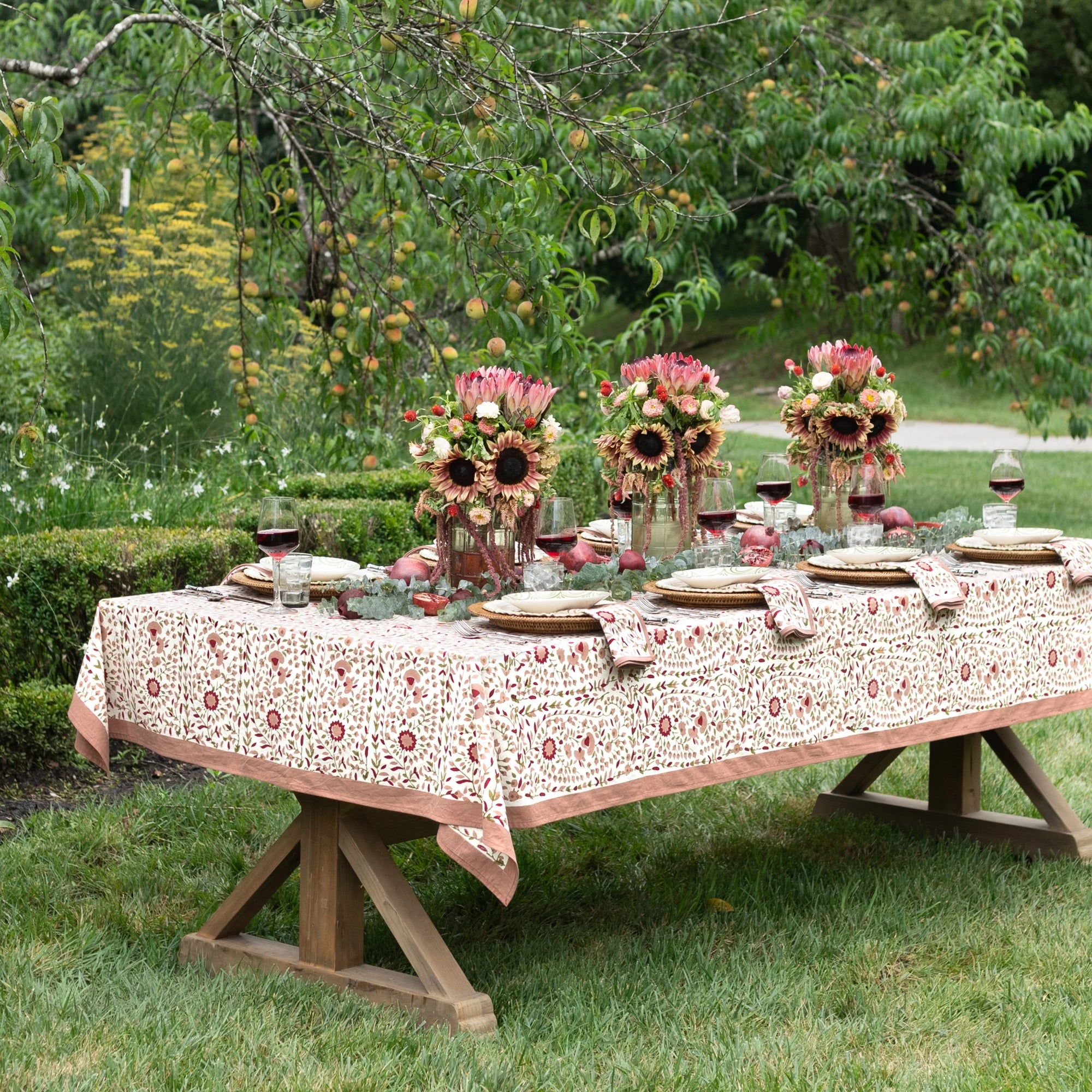 A rectangular wooden table set outdoors on grass with a beige tablecloth featuring a red and pink floral pattern. The table is styled with sunflower centerpieces, plates, folded napkins, and wine glasses, surrounded by green garden foliage.