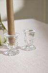 Two clear glass candle holders on a white surface with a brown candle in one of them.