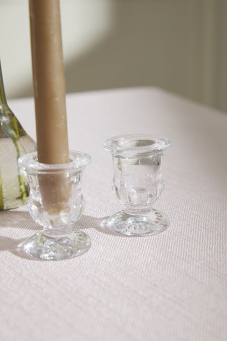 Two clear glass candle holders on a white surface with a brown candle in one of them.