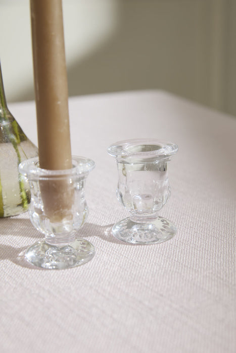 Two clear glass candle holders on a white surface with a brown candle in one of them.