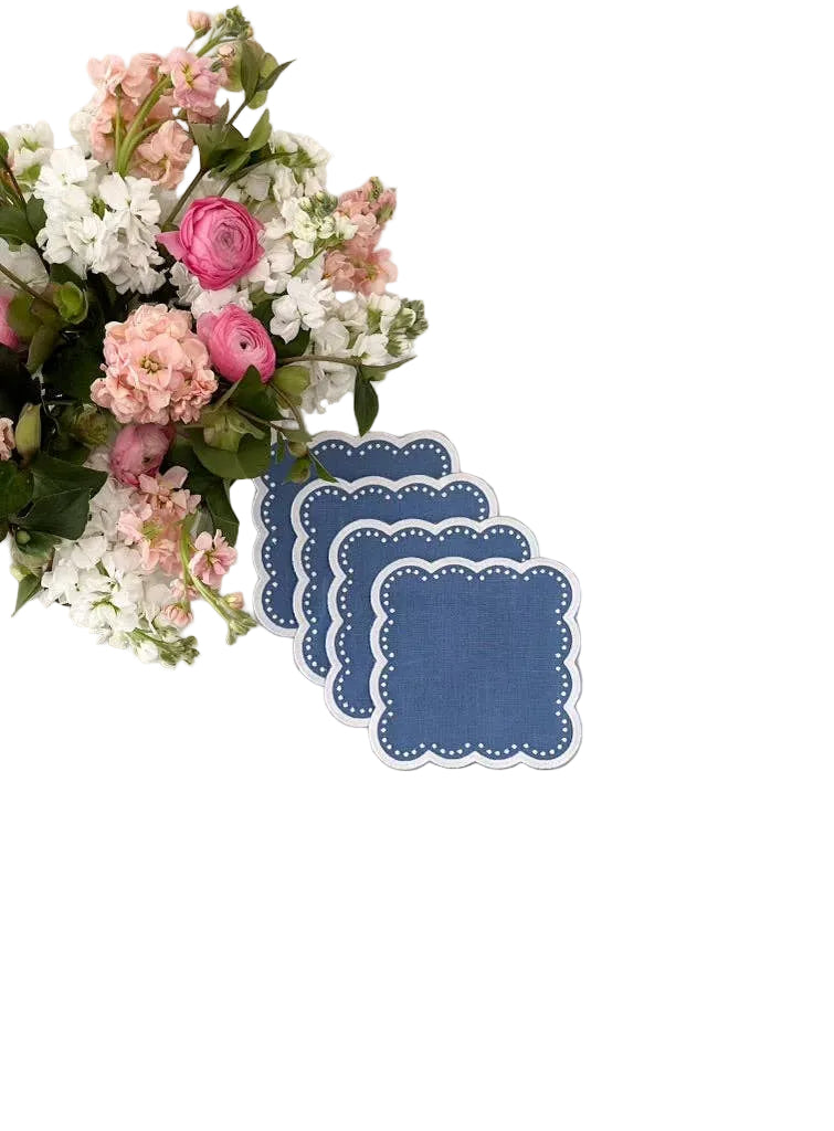 A set of four blue scalloped cocktail napkins placed on a white surface, with a bouquet of multicolored flowers in the background.