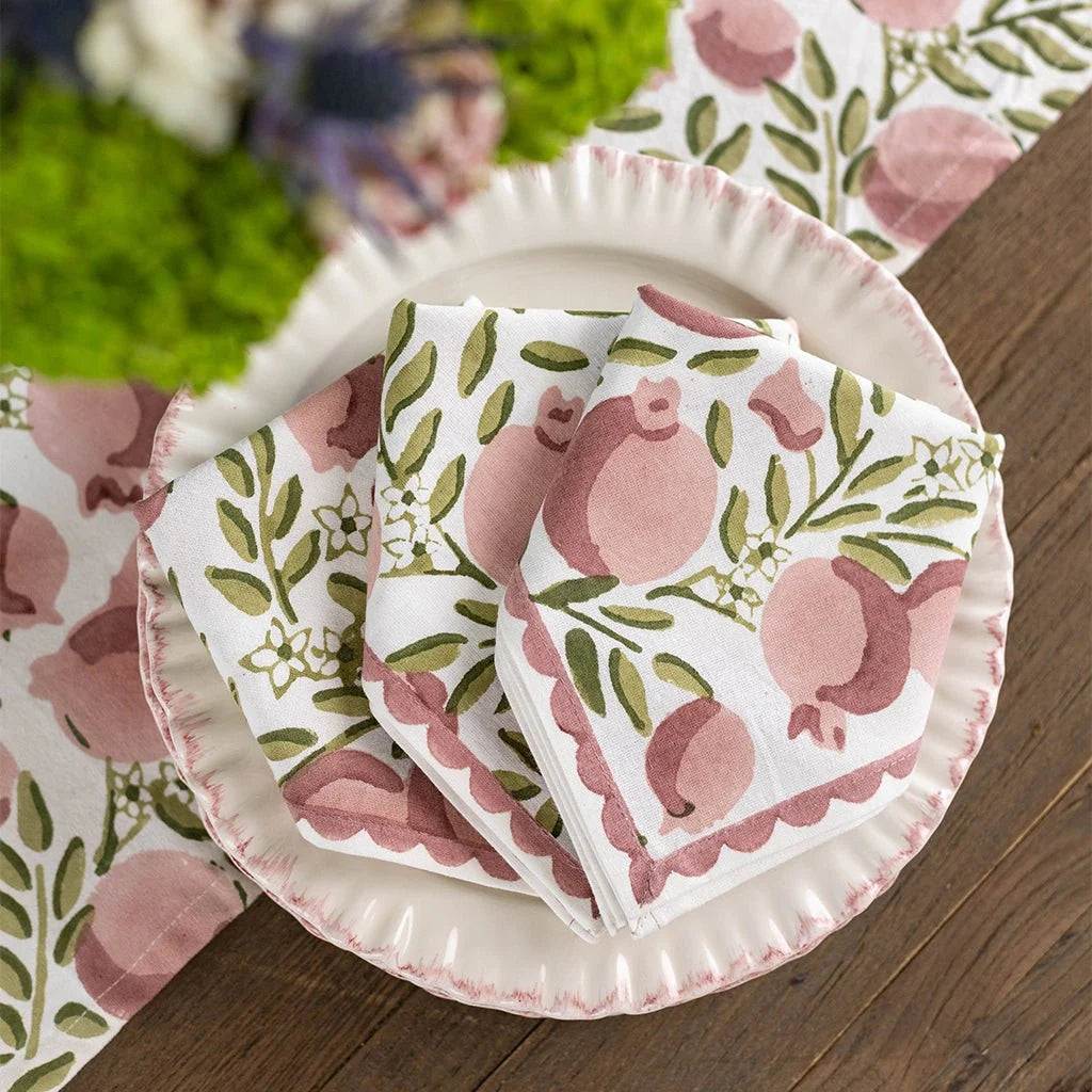 A stack of white paper napkins with a pink pomegranate and green leaf pattern, arranged on a matching white paper plate with pink scalloped edges on a wooden table.
