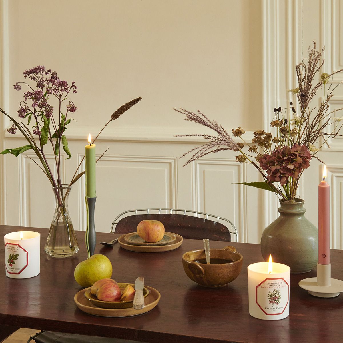 Dining table setting with candles, fruit, and decorative vases in a room with white paneled walls.